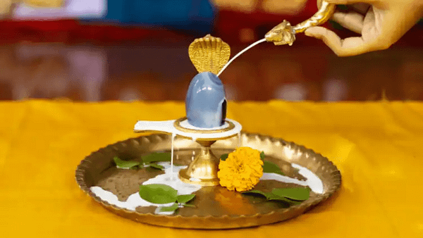 A hand performing the rudrabhishek ritual by pouring milk on a shiva lingam adorned with marigold flowers and bilva leaves on a brass plate.